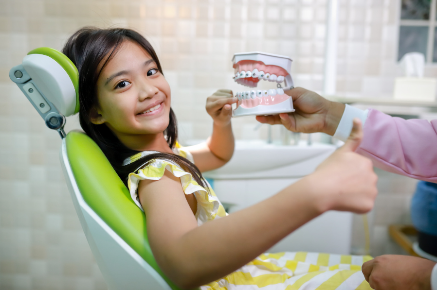 Child in dental chair looking at braces model for early orthodontics.